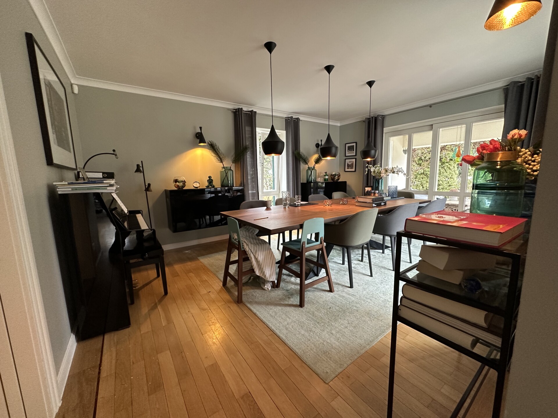 Das Bild zeigt ein Foto von einem Esszimmer mit Holzboden, bodentiefen fenstern zum Garten hin, einem großen Esstisch mit Esszimmerstühlen für 10 Personen, einem Klavier und gestrichenen Wänden in einem grünen Farbton.